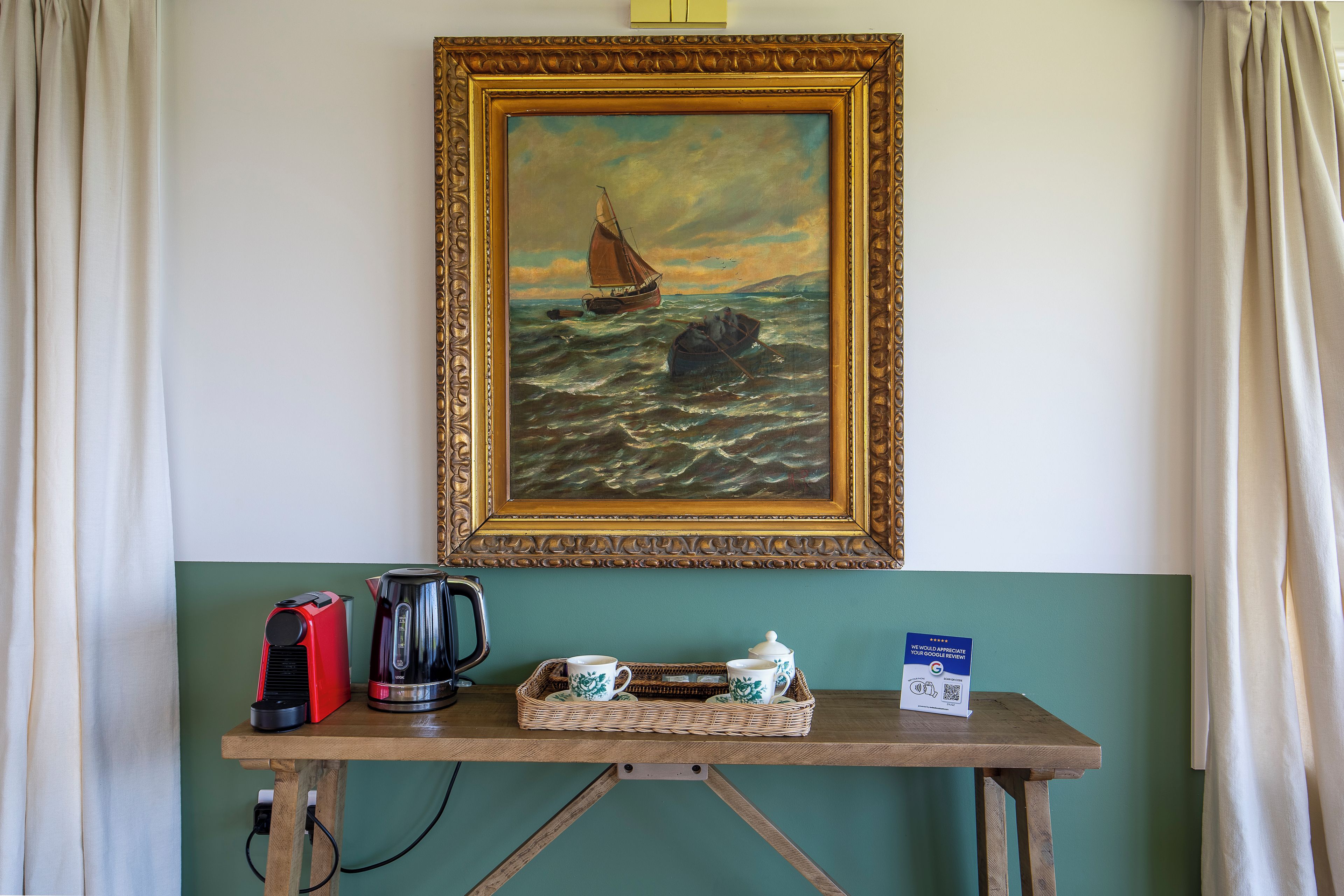 A painting of a sailboat on rough seas, a coffee maker, a kettle, and a tea set on a wooden table against a teal wall.