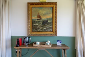 A painting of a sailboat on rough seas, a coffee maker, a kettle, and a tea set on a wooden table against a teal wall.