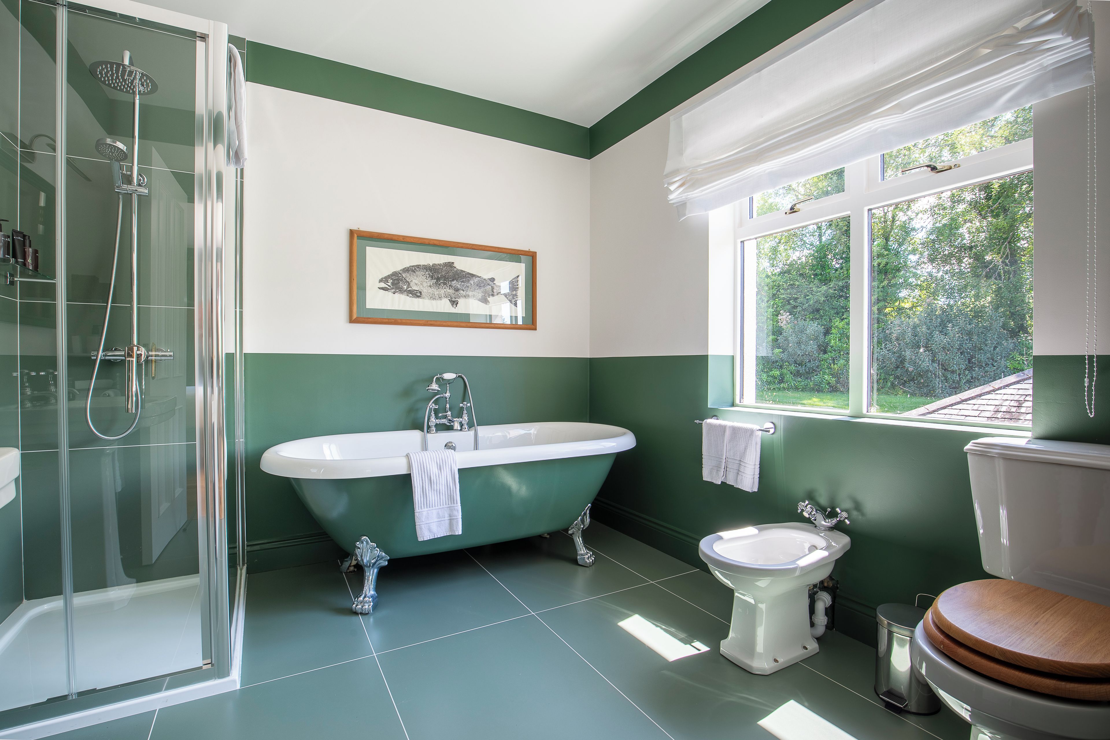 A bathroom with a glass-enclosed shower, a freestanding bathtub, a toilet with a wooden seat, and a window with a view of greenery.