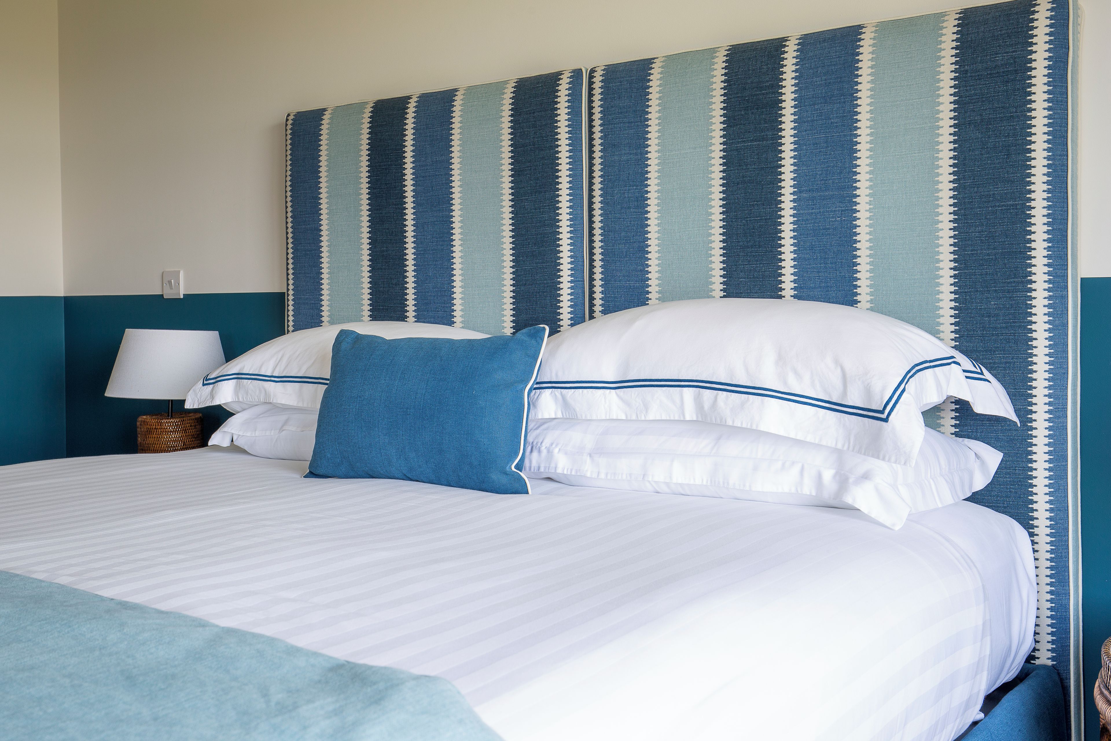 Een net opgemaakt bed met witte lakens en blauwe accenten, een bedlampje en een gestreepte muur op de achtergrond.