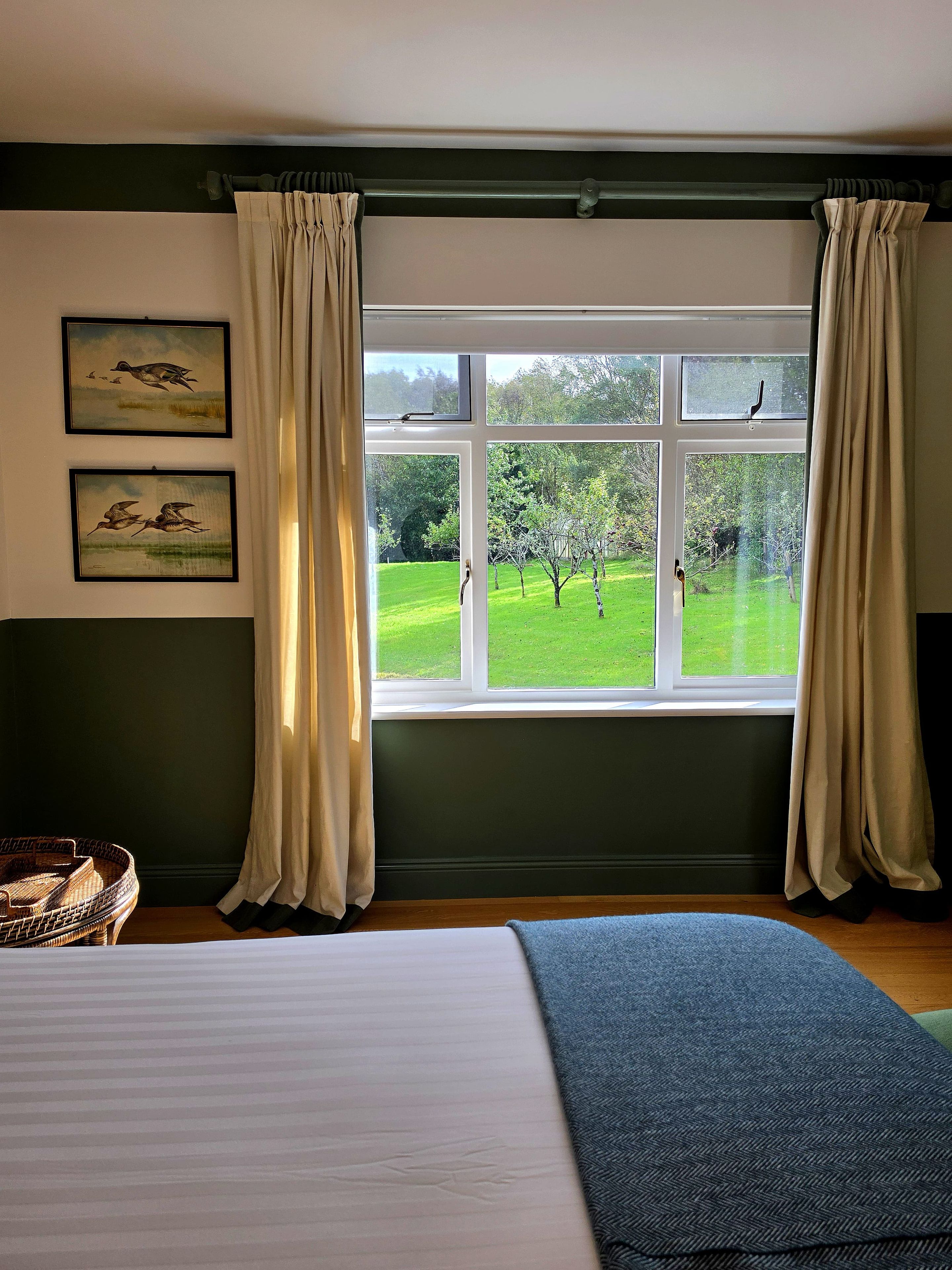 A bedroom with a large window, two framed pictures on the wall, a bed, and a wicker chair.