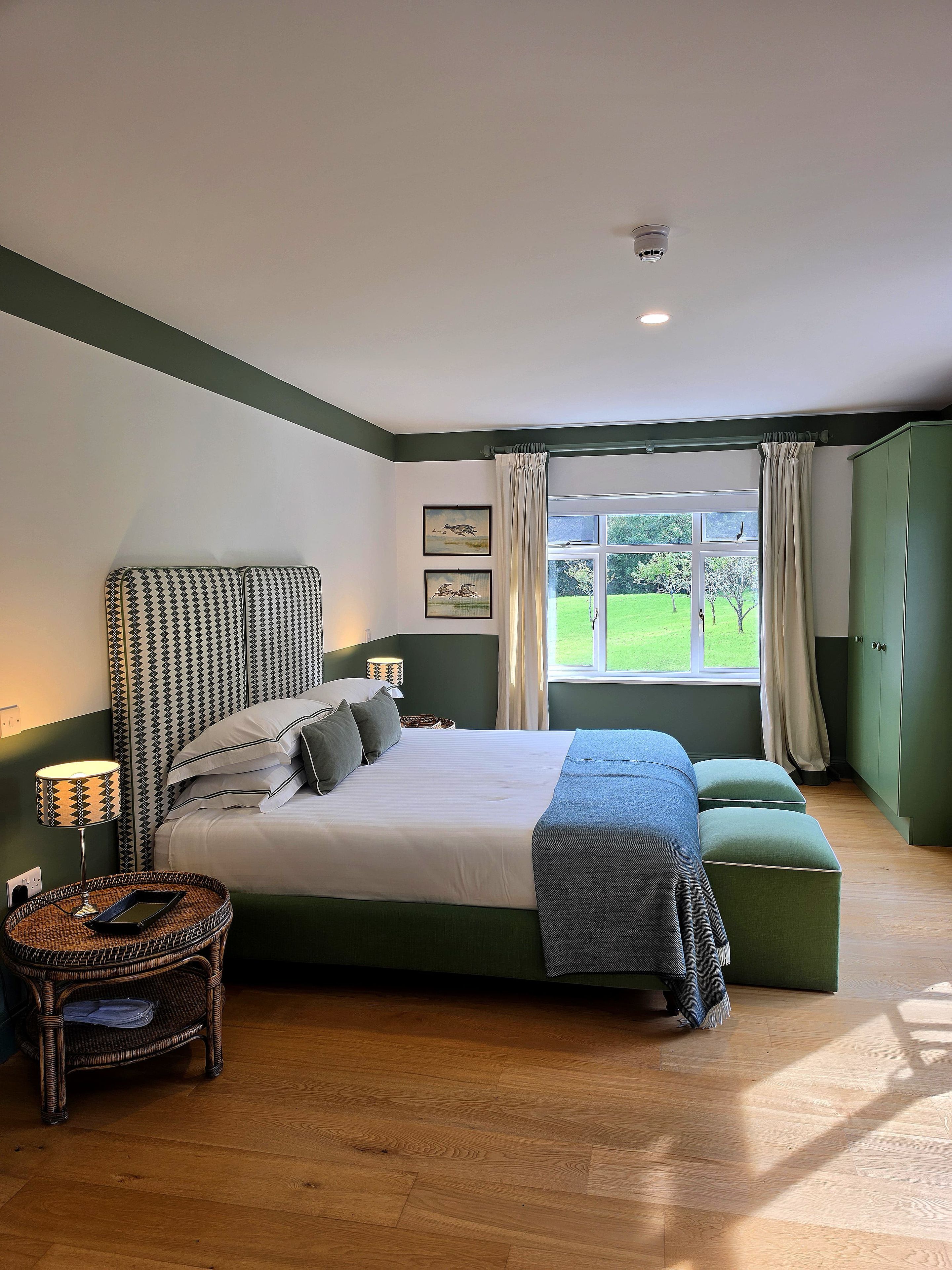 Un dormitorio con una cama grande, una mesita de noche con lámpara, una ventana con cortinas y vistas a un paisaje verde.