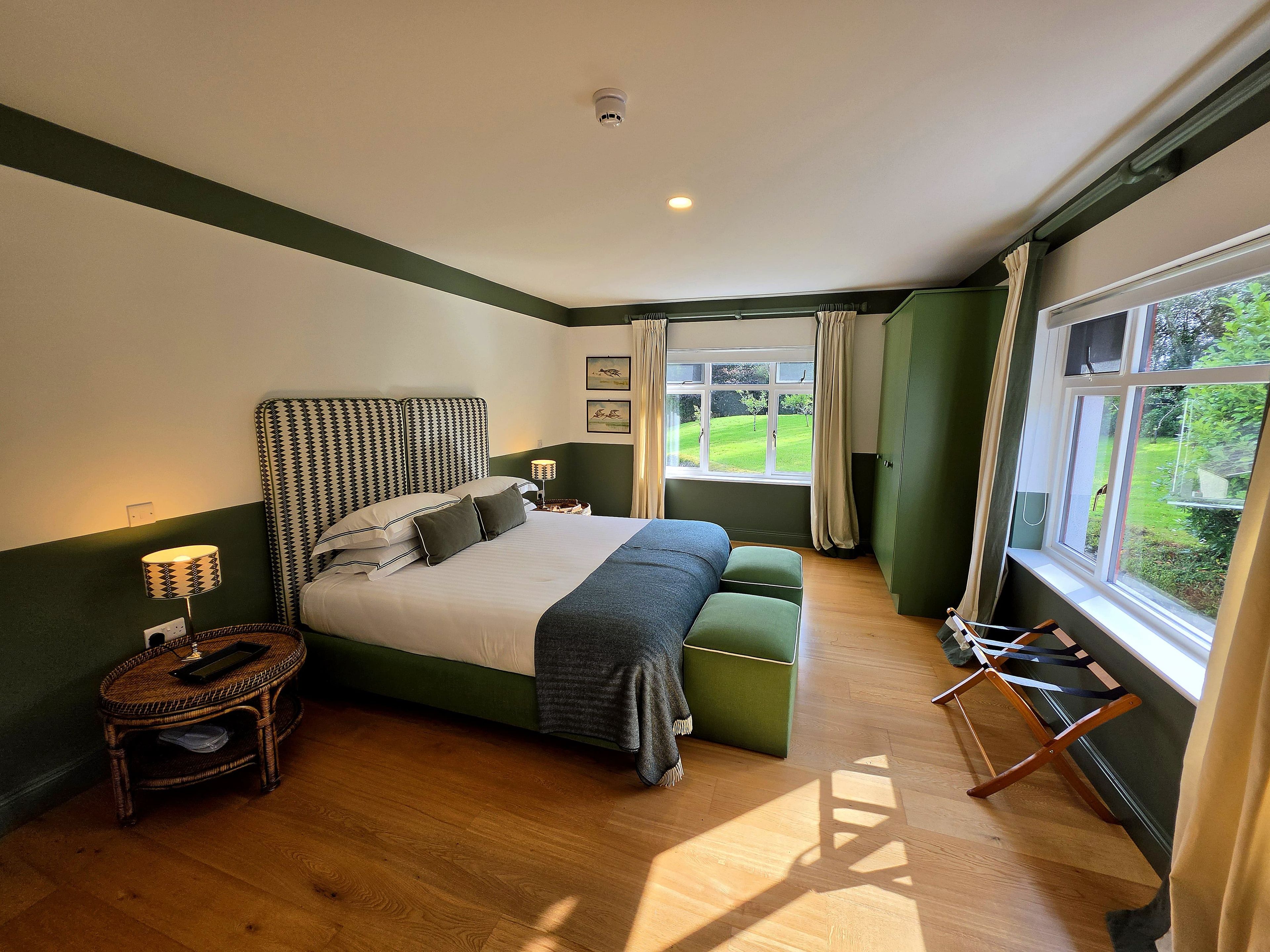Un dormitorio con una cama grande, una mesita de noche con lámpara, un banco verde y vistas a un jardín por una ventana.