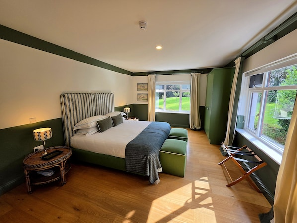A bedroom with a large bed, a bedside table with a lamp, a green bench, and a view of a garden through a window.
