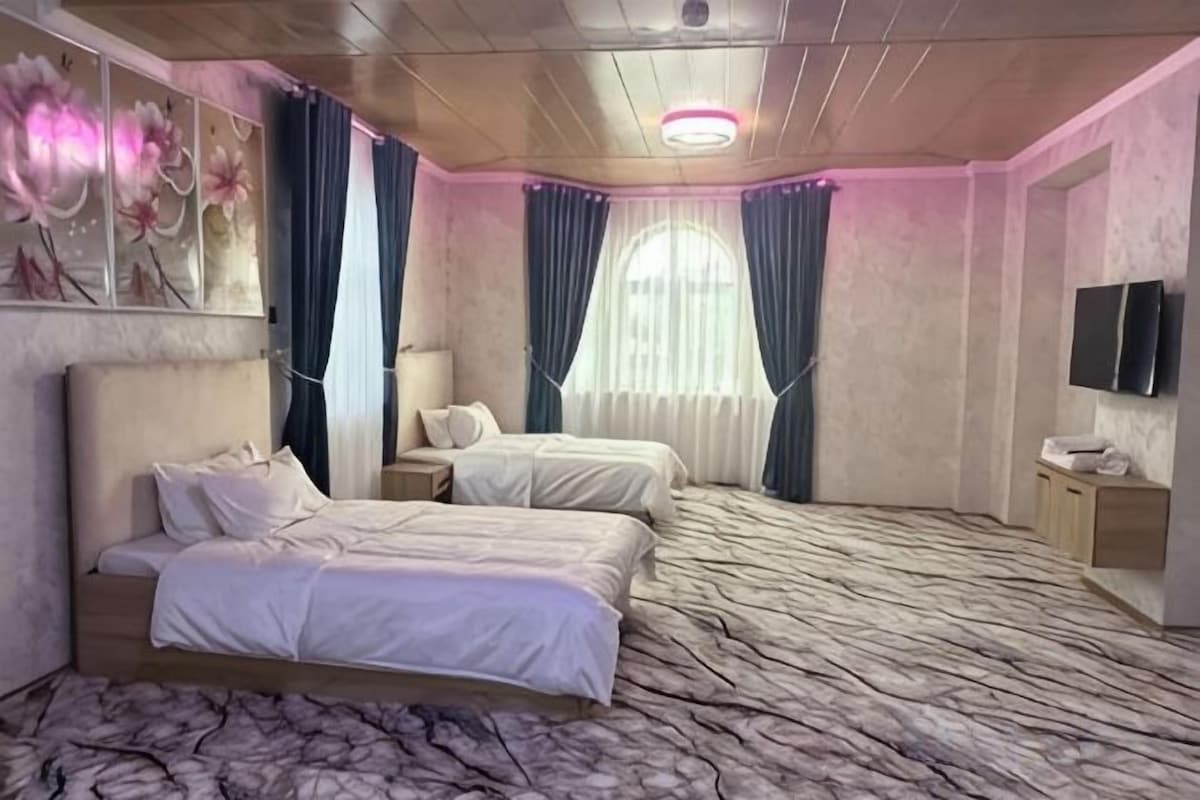 A hotel room with two beds, a marble floor, a large window with curtains, a flat-screen TV, and a wall-mounted TV.