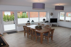 A dining area with a wooden table and chairs, a television, and large windows with blinds. - Holiday home with 4 bedrooms (Norddeich)