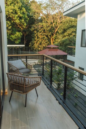 A balcony with a wicker chair and a small table, a metal railing, and a view of trees and a building. - Howler Monkey Loft (Playas Del Coco)