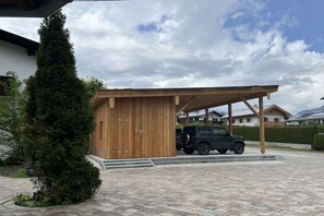 A covered carport with a vehicle parked inside, a wooden building with a sloped roof, and a paved driveway. - Holiday apartment with 2 bedrooms (Bischofswiesen)