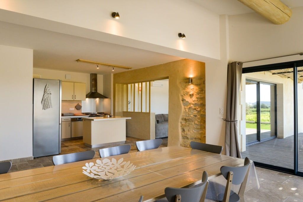 A modern kitchen with a central island, a dining area with a wooden table and chairs, and a view through a glass door to an outdoor deck.