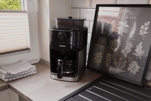 Cozinha com cafeteira, ilustração emoldurada de um jardim de ervas e janela com persianas.