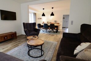 A modern living room with a sofa, a coffee table, and a TV mounted on the wall. There is a dining area with a table and chairs in the background. - Holiday apartment in Lütetsburg with sauna (Lütetsburg)