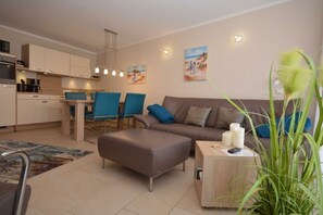 Een moderne woonkamer met een bank, een voetenbankje en een salontafel. Er is een eethoek met stoelen en een tafel. De kamer heeft een keuken met kastjes en een fornuis.