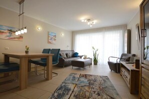 A modern living room with a dining area, a sofa set, a coffee table, and a large window with curtains. - Holiday apartment with 2 bedrooms (Norddeich)