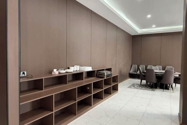 A modern dining area with a long table and chairs, a built-in shelving unit with various items, and a tiled floor.