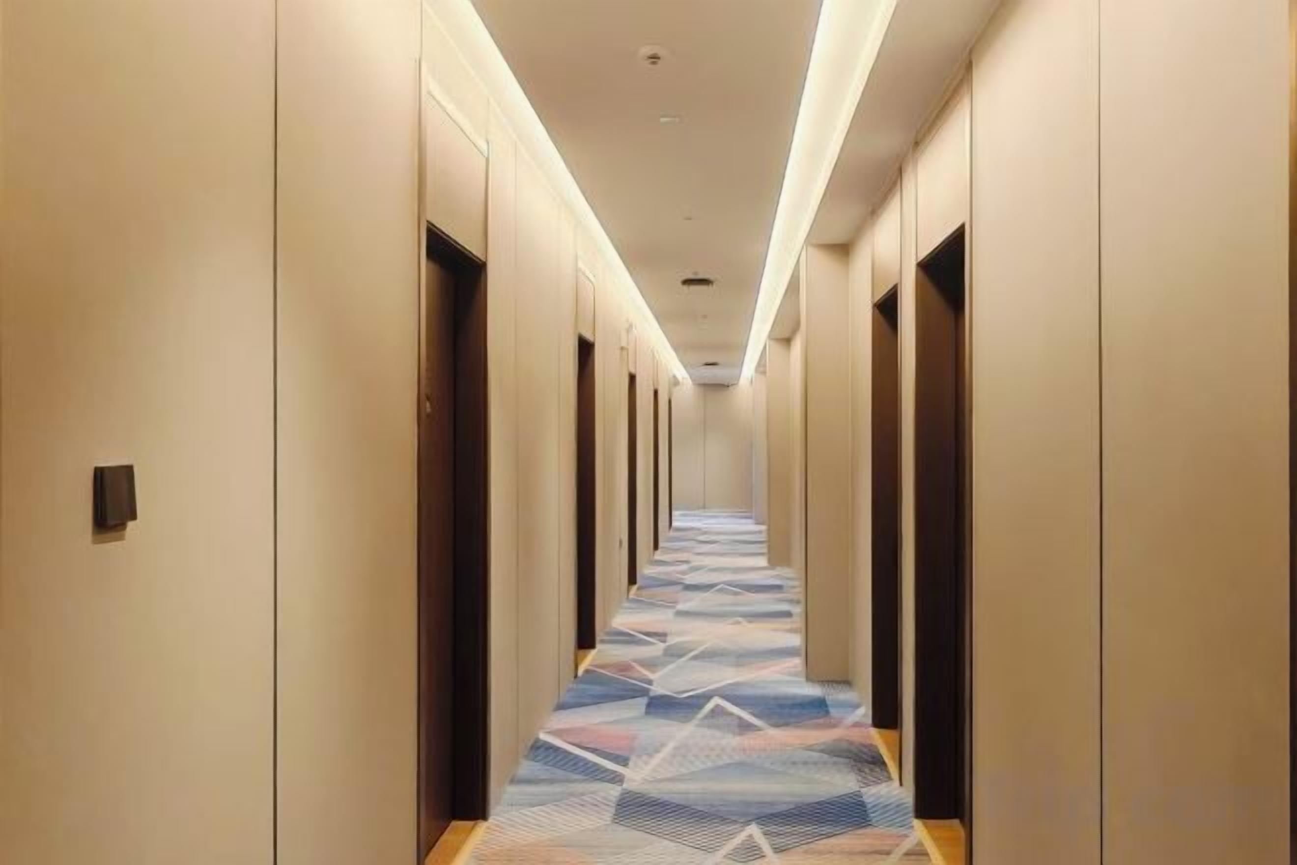 A hotel hallway with doors on both sides, a patterned carpet, and a ceiling with recessed lighting.
