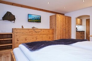 A bedroom with a wooden headboard, a bed with white bedding, a dark throw blanket, a mounted picture of a mountain landscape, and a backpack on the wall. - Holiday apartment no. 3 - Guesthouse zum Wurm (Reit im Winkl)