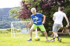 Un champ d’herbe avec un but de football, deux personnes qui jouent au football et une table de pique-nique en arrière-plan.