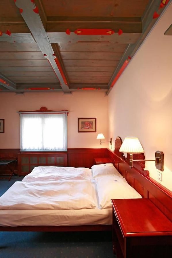 Una habitación de hotel con cama, escritorio, silla, ventana con cortinas y lámpara fijada a la pared.