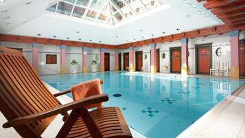 Una piscina interior con sillones de madera, techo de cristal y suelo de baldosas.