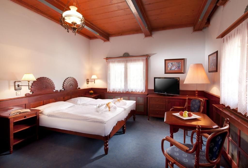 A hotel room with a wooden bed, bedside tables, a television, a desk with a chair, and a window with curtains.