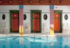 Una piscina con agua cristalina, rodeada de sillas blancas y una fila de puertas numeradas del 18 al 20.