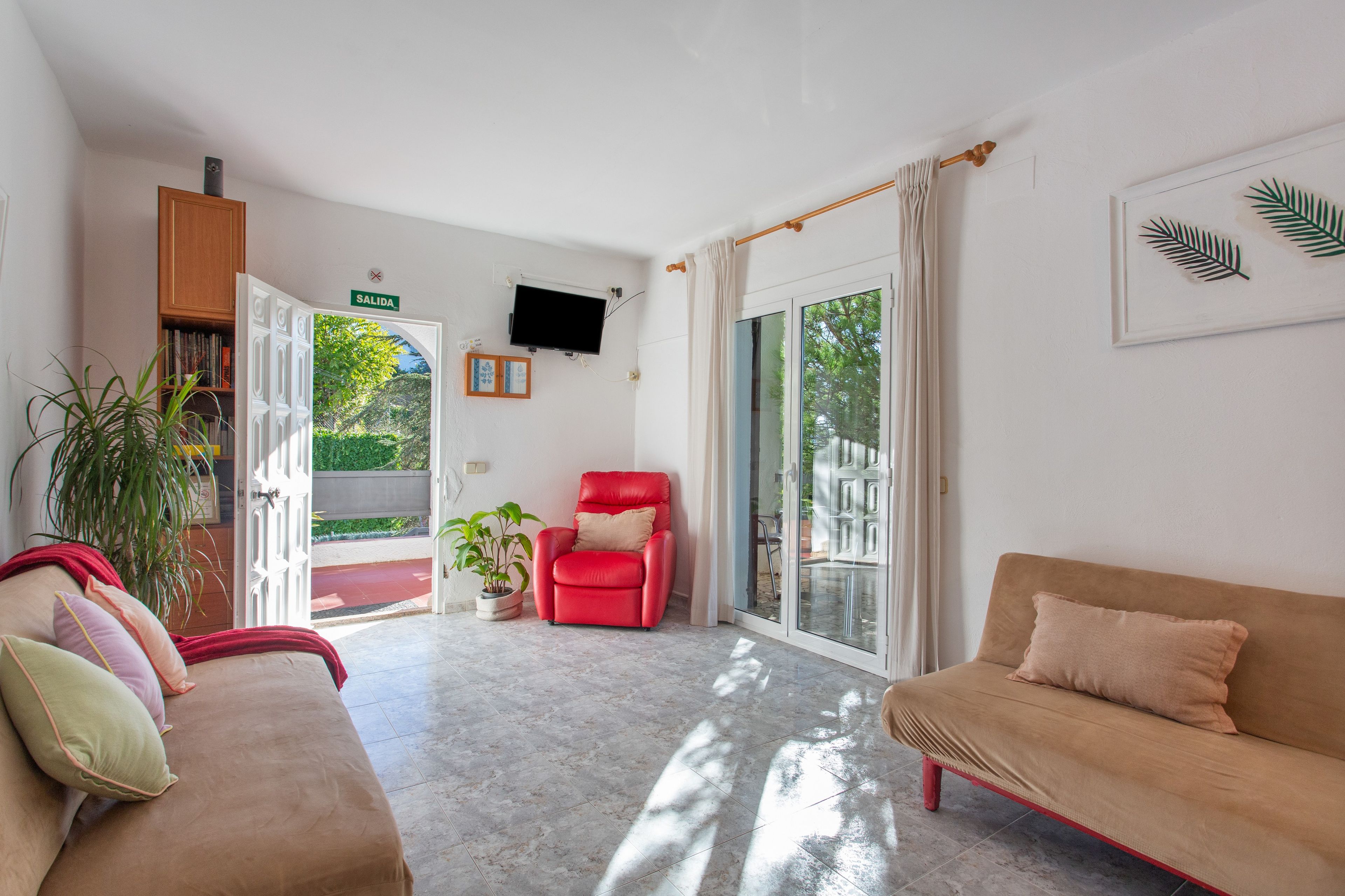 Un amplio salón con una silla roja, un sofá beige y un televisor montado en la pared.