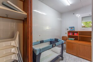 A room with a crib, a wooden cabinet with books, and a window with blinds.