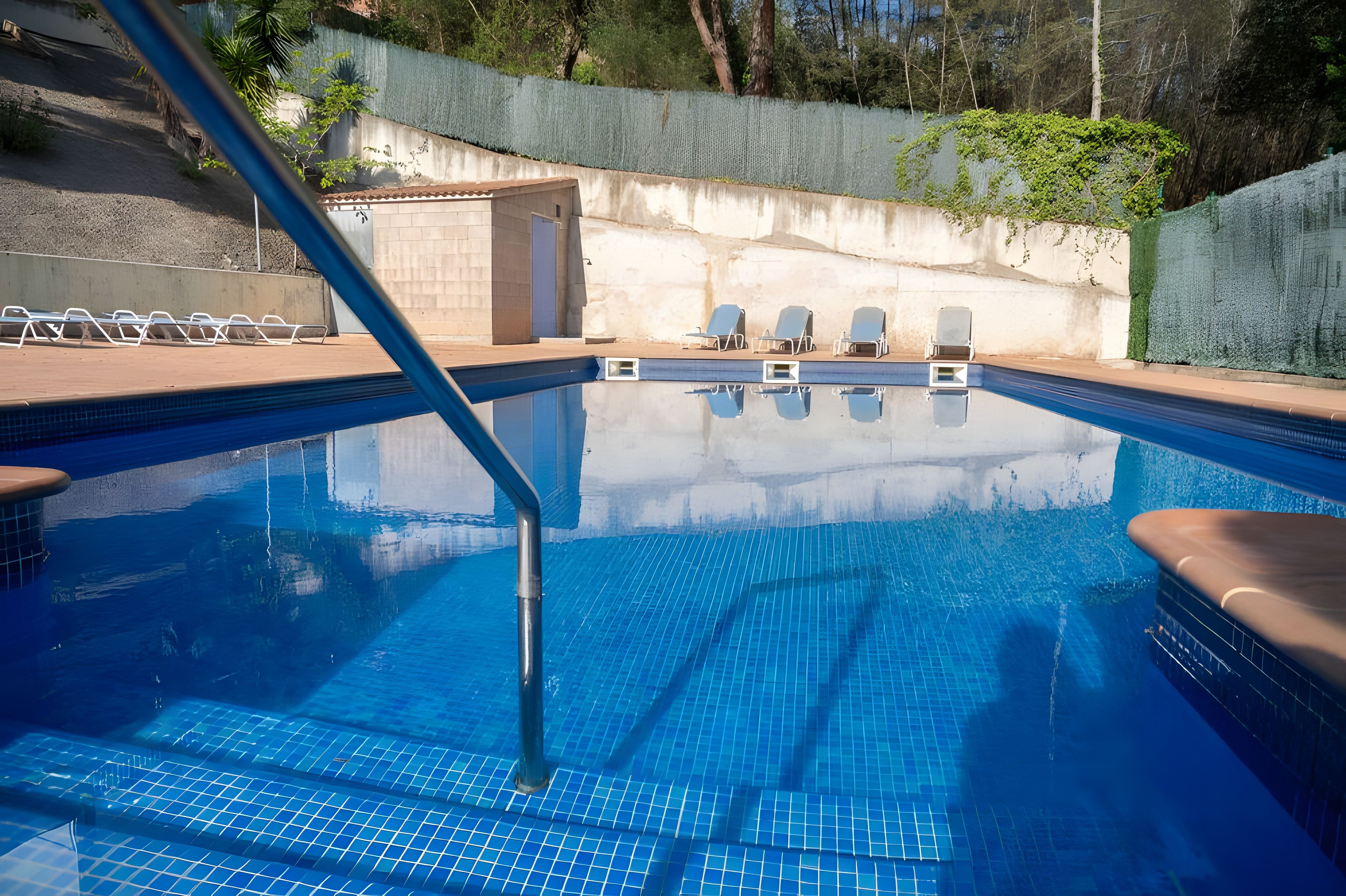 Una piscina con agua cristalina azul, rodeada de sillas de descanso y un muro de hormigón con plantas.
