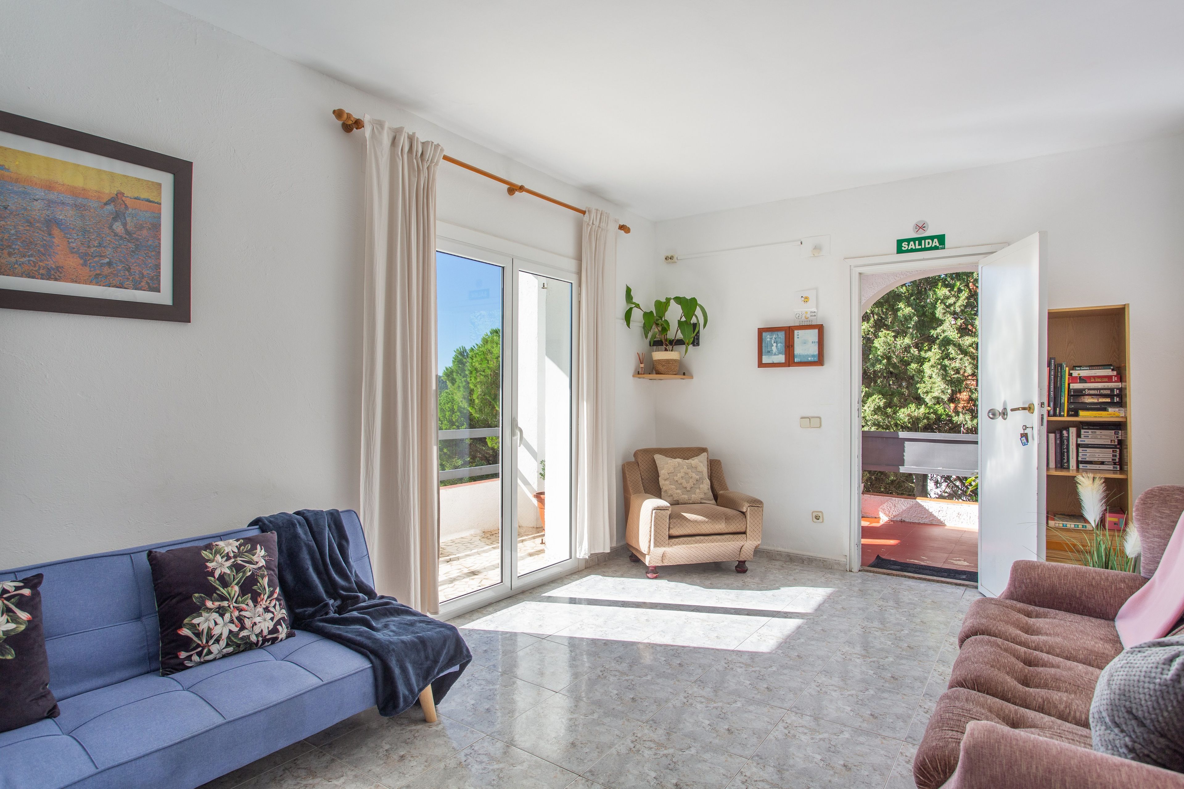A living room with a blue sofa, a beige armchair, a bookshelf, and a door leading to an outdoor area.