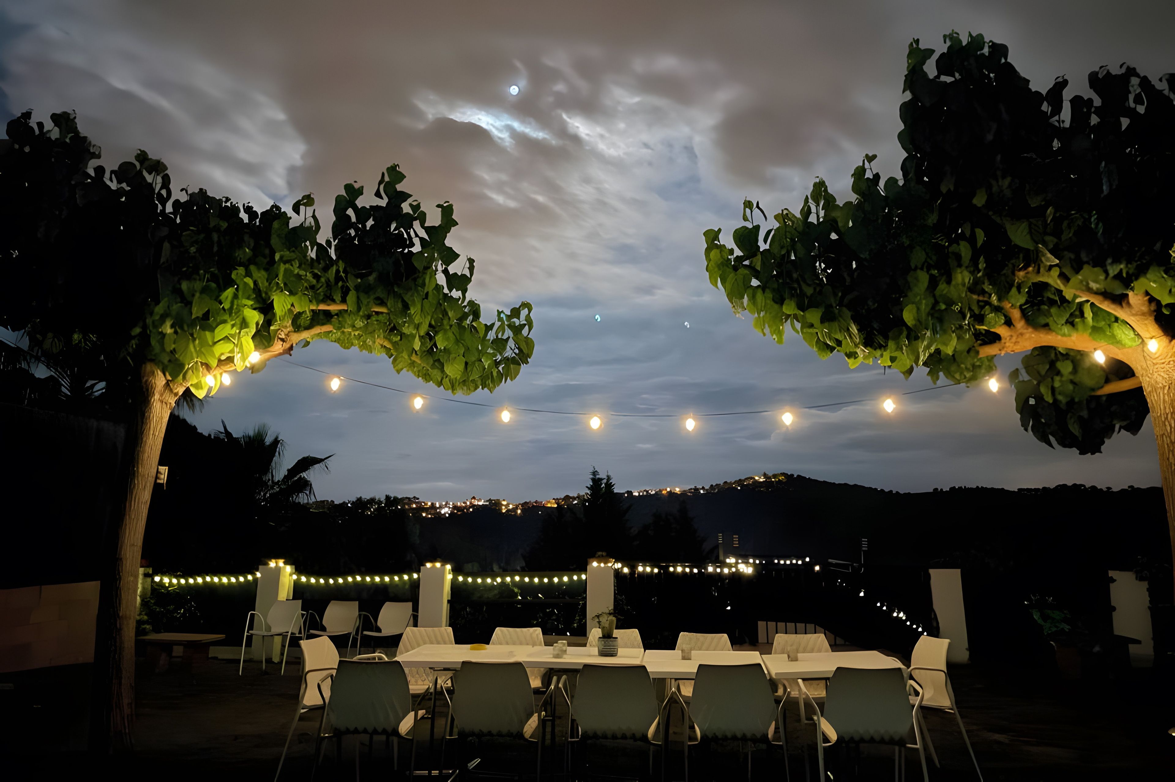 Un área de comedor al aire libre con mesas y sillas bajo una pérgola, luces colgantes y vistas a un pueblo lejano.