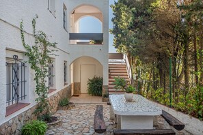 A courtyard with a stone floor, a wooden bench, a potted plant, and a white wall with a window and door.