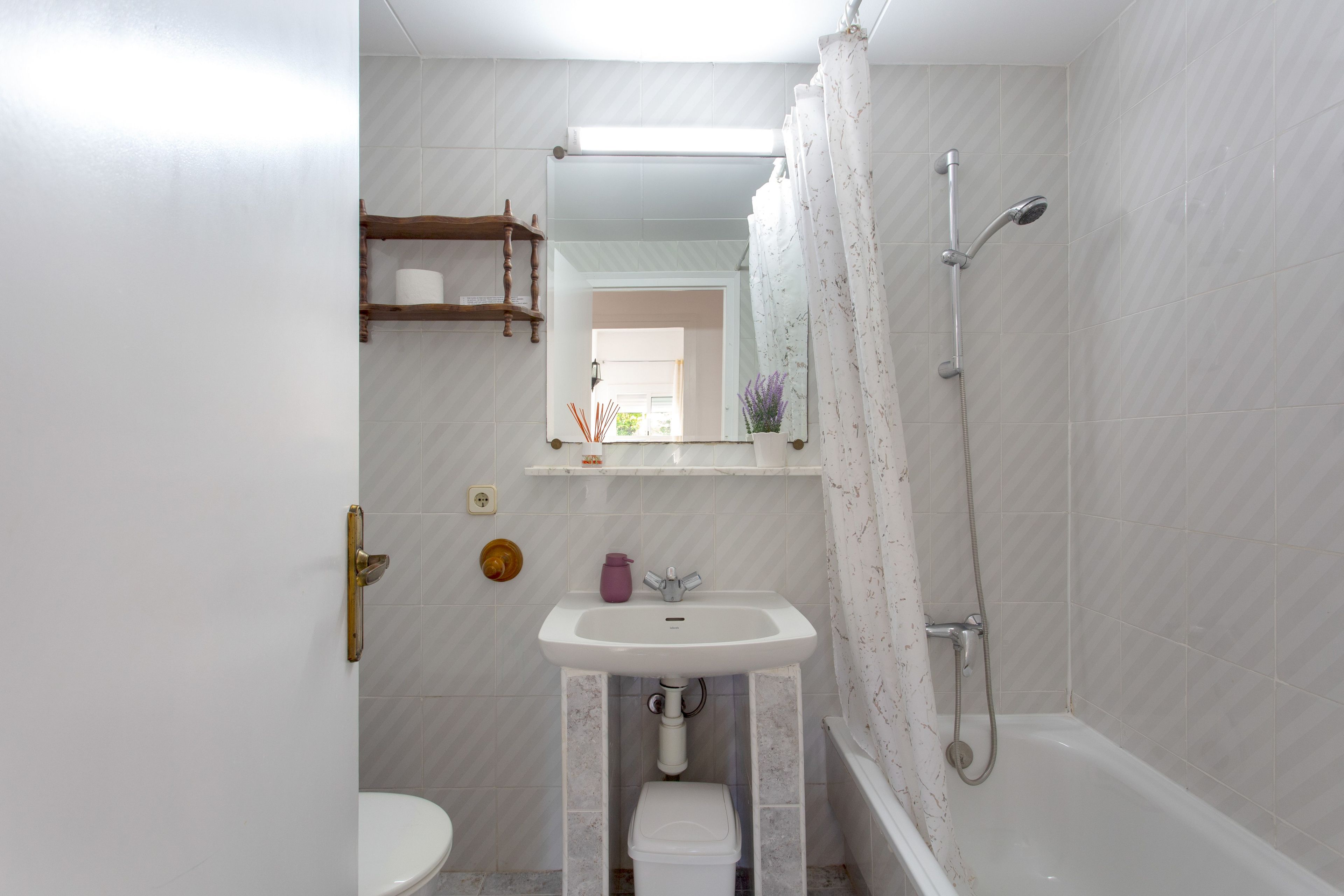 Un baño con inodoro, lavabo y ducha. Hay un espejo sobre el lavabo y un estante con un rollo de papel higiénico.