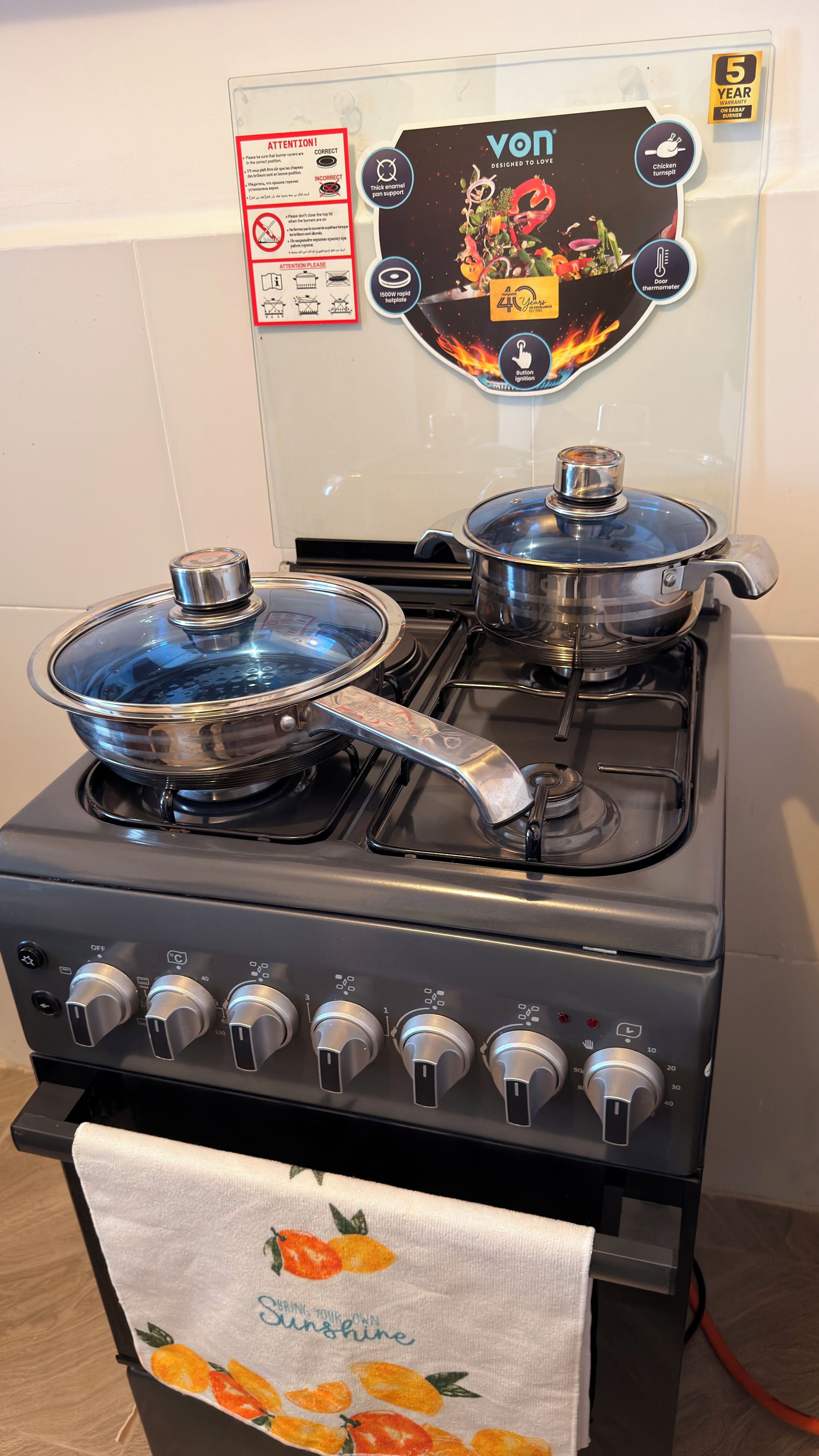 Une cuisine avec deux casseroles en acier inoxydable sur une cuisinière, un autocollant indiquant une garantie de cinq ans et un panneau d’avertissement de sécurité.