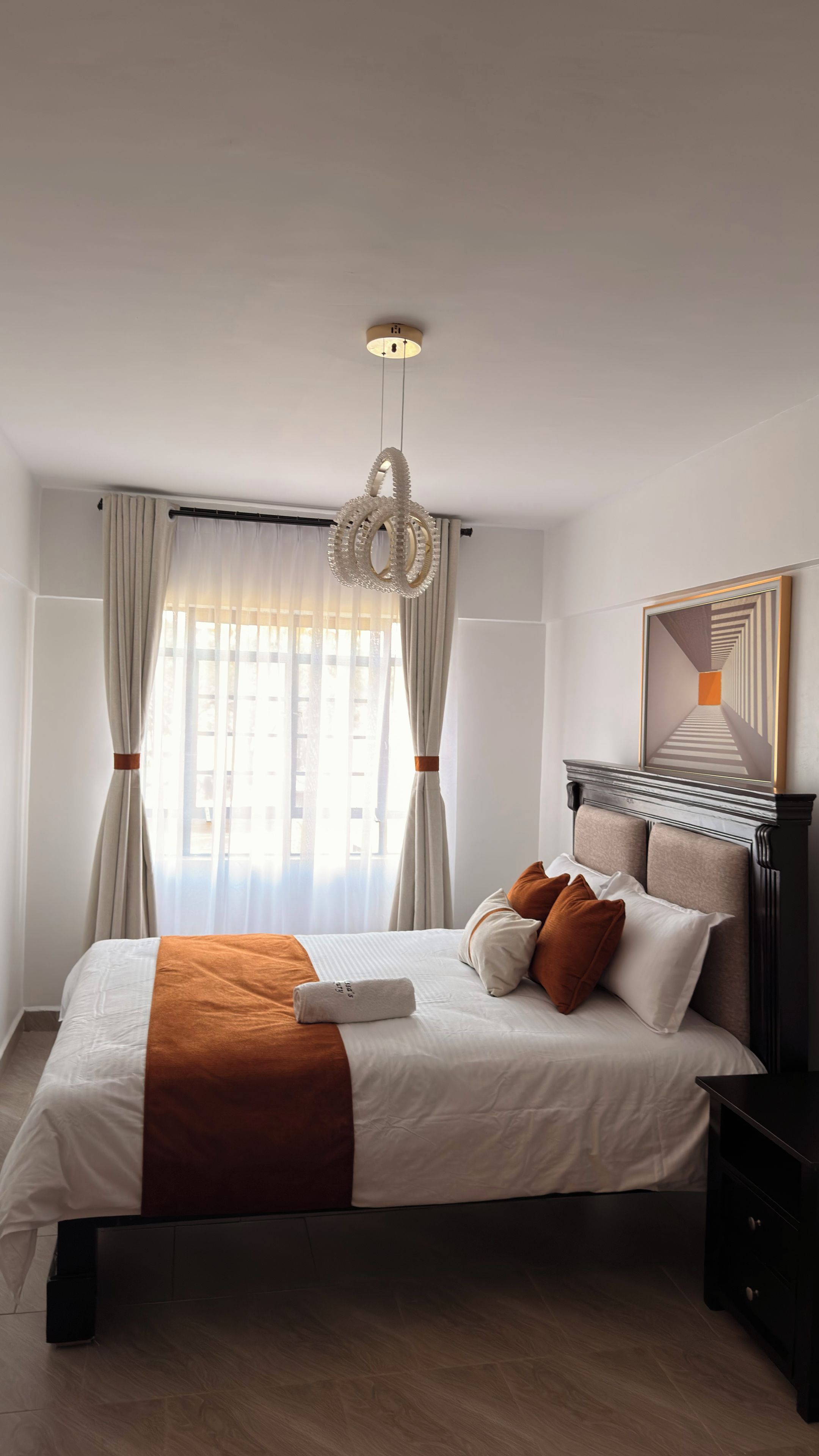 Ein Hotelzimmer mit einem großen Bett, einer Hängelampe, einem Fenster mit Vorhängen und einem gerahmten Kunstwerk an der Wand.