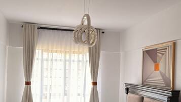 Ein Hotelzimmer mit einem großen Bett, einer Hängelampe, einem Fenster mit Vorhängen und einem gerahmten Kunstwerk an der Wand.