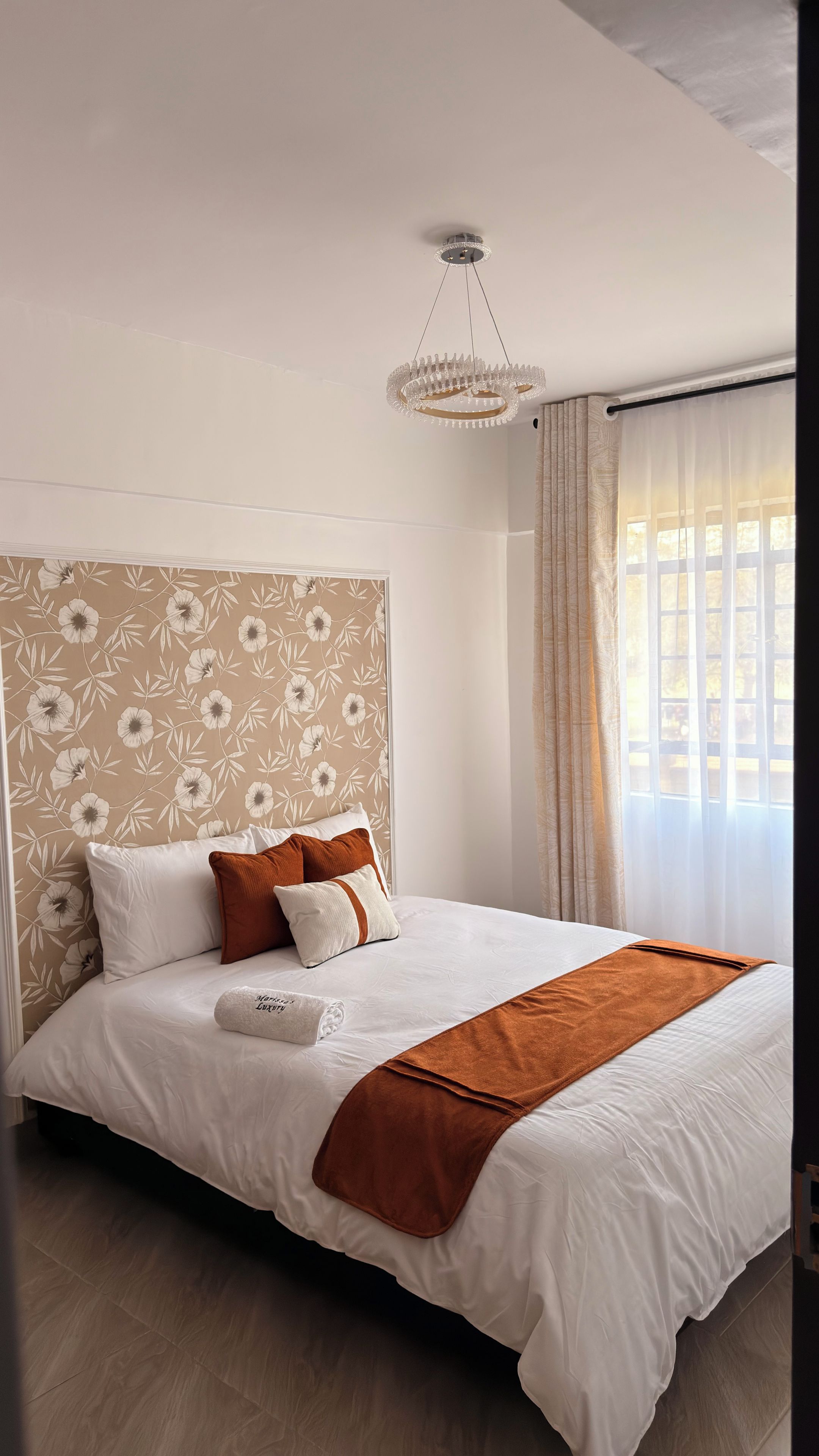 A hotel room with a large bed, a chandelier, floral wallpaper, and a window with curtains.