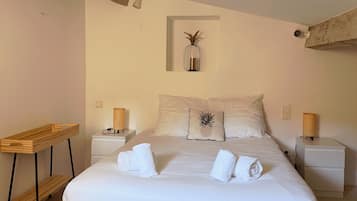 Een net opgemaakt bed met witte lakens en een kussen in de vorm van een ananas, een klein tafeltje met een rieten mand en een wandlamp.