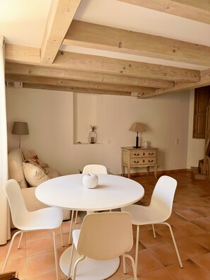 Una habitación con una mesa redonda blanca rodeada de cuatro sillas blancas, un sofá con cojines, un mueble pequeño y una lámpara en la pared.