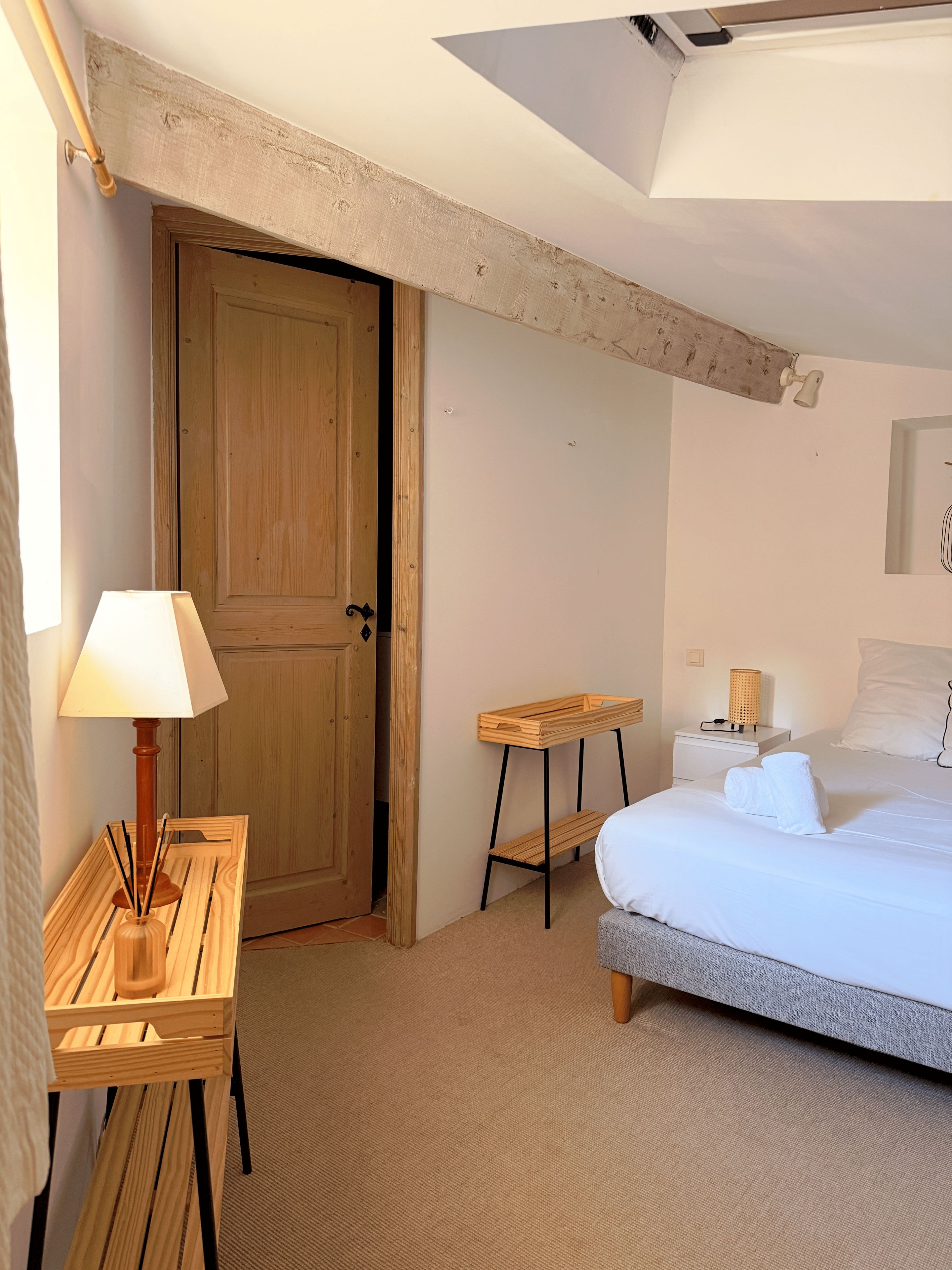 Une chambre avec une porte en bois, une table de chevet avec une lampe, un petit tabouret en bois et un lit avec une literie blanche.