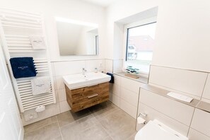 Ein modernes Badezimmer mit einem weißen Waschtisch, einem Spiegel über dem Waschbecken und einem Fenster für natürliches Licht.