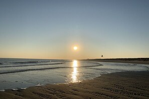 Een strand met een heldere lucht, de zon die in het water weerspiegelt, en voetstappen in het zand.