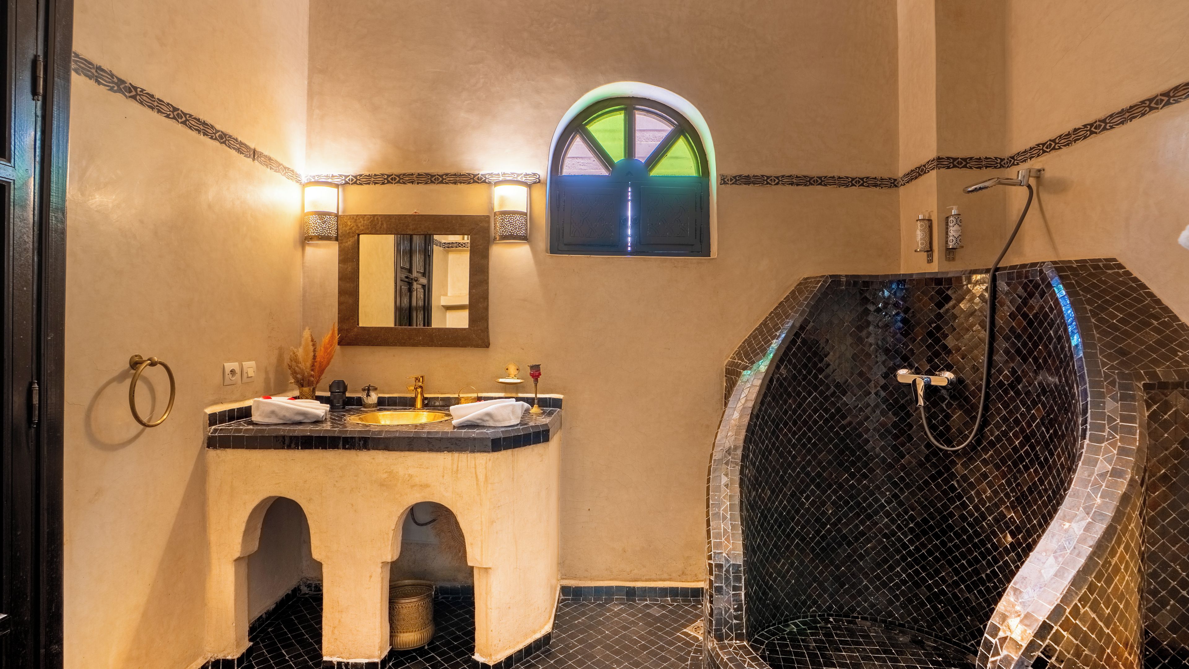 Ein Badezimmer mit Waschtisch, Spiegel, Dusche mit Mosaikfliesen und einem kleinen Fenster mit Blick auf Grün.