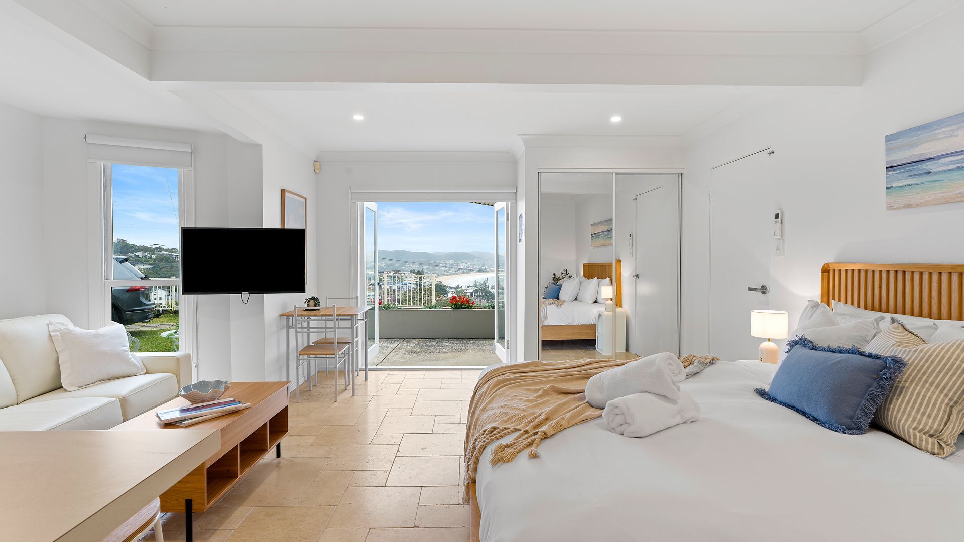 A hotel room with a bed, a desk, a TV, and a balcony with a view of the city.