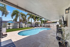 A curved swimming pool with a covered patio area, surrounded by palm trees and a white fence. - Sun, Pool & Sand- Fort Lauderdale Style Escape! (Fort Lauderdale)