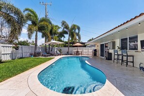 A swimming pool with a curved edge, surrounded by a white fence, palm trees, and a patio area with chairs and umbrellas. - Sun, Pool & Sand- Fort Lauderdale Style Escape! (Fort Lauderdale)