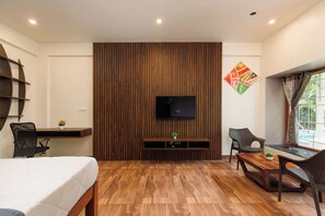 A hotel room with a wooden floor, a bed, a desk, a TV mounted on a wooden panel, and a window with curtains. - Bougainvillea by Stayjade Poolbonfire Mysore Rd (Channapatna)