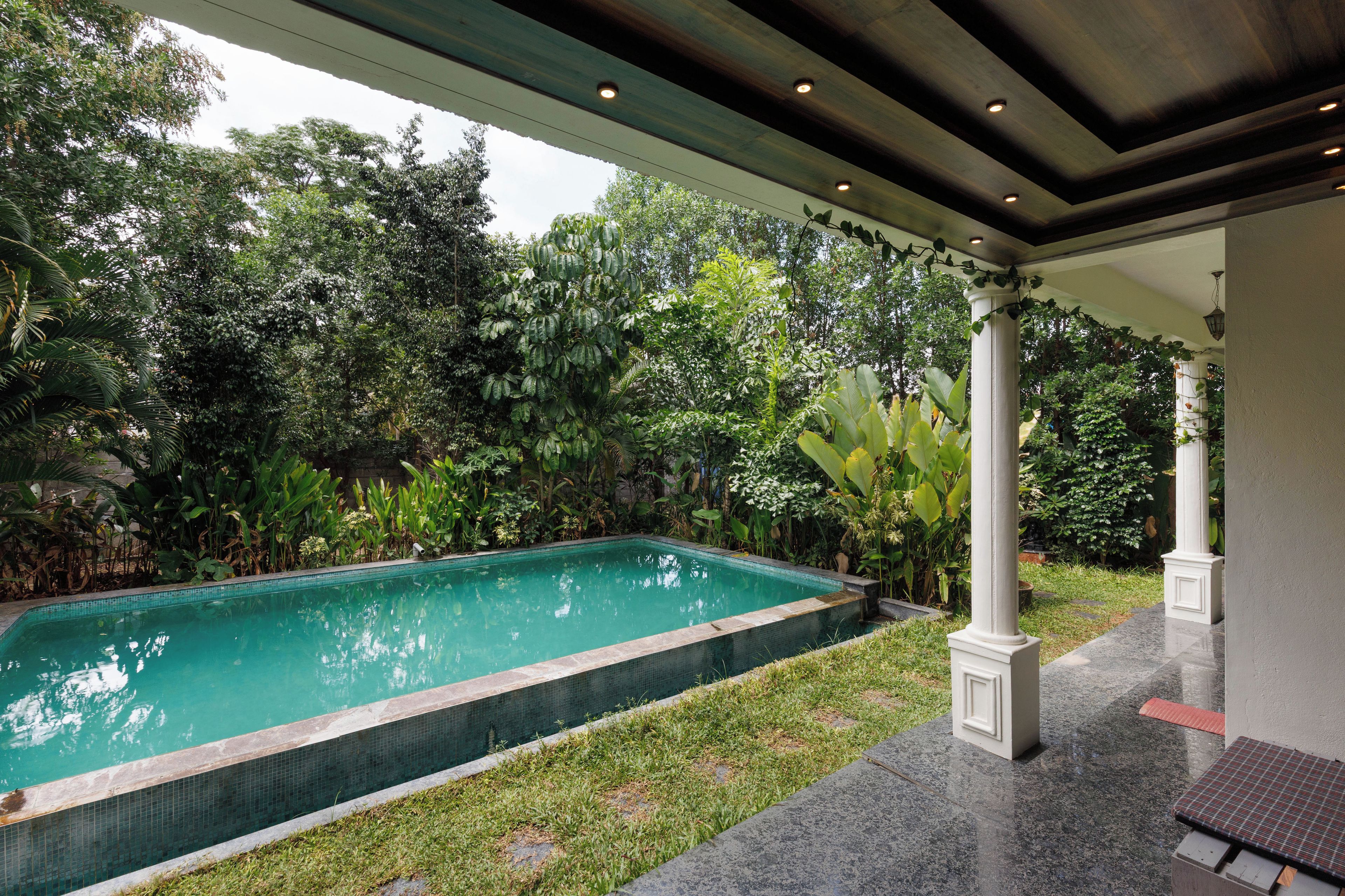 A swimming pool with a tiled edge, surrounded by lush greenery and a covered patio area with columns.