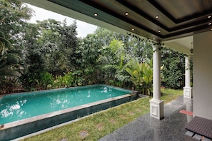 Une piscine dont le bord est revêtu de carrelage, entourée d’une végétation luxuriante et dotée d’un patio couvert avec des colonnes.
