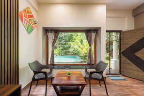 A room with a wooden floor, two wicker chairs, a wooden table, a window with curtains, and a view of a swimming pool. - Bougainvillea by Stayjade Poolbonfire Mysore Rd (Channapatna)