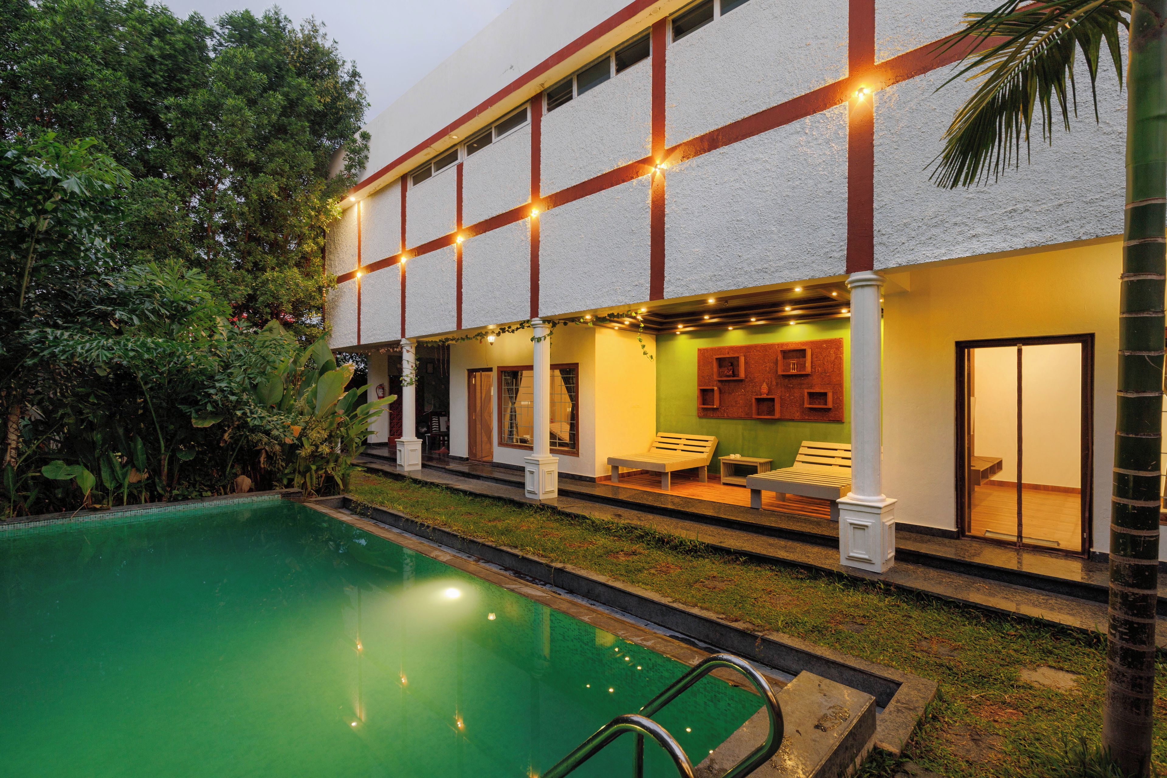 Un espace aménagé au bord de la piscine, avec une piscine à eau claire, un coin salon couvert avec des chaises, et un bâtiment à la façade blanche agrémentée de touches de rouge.